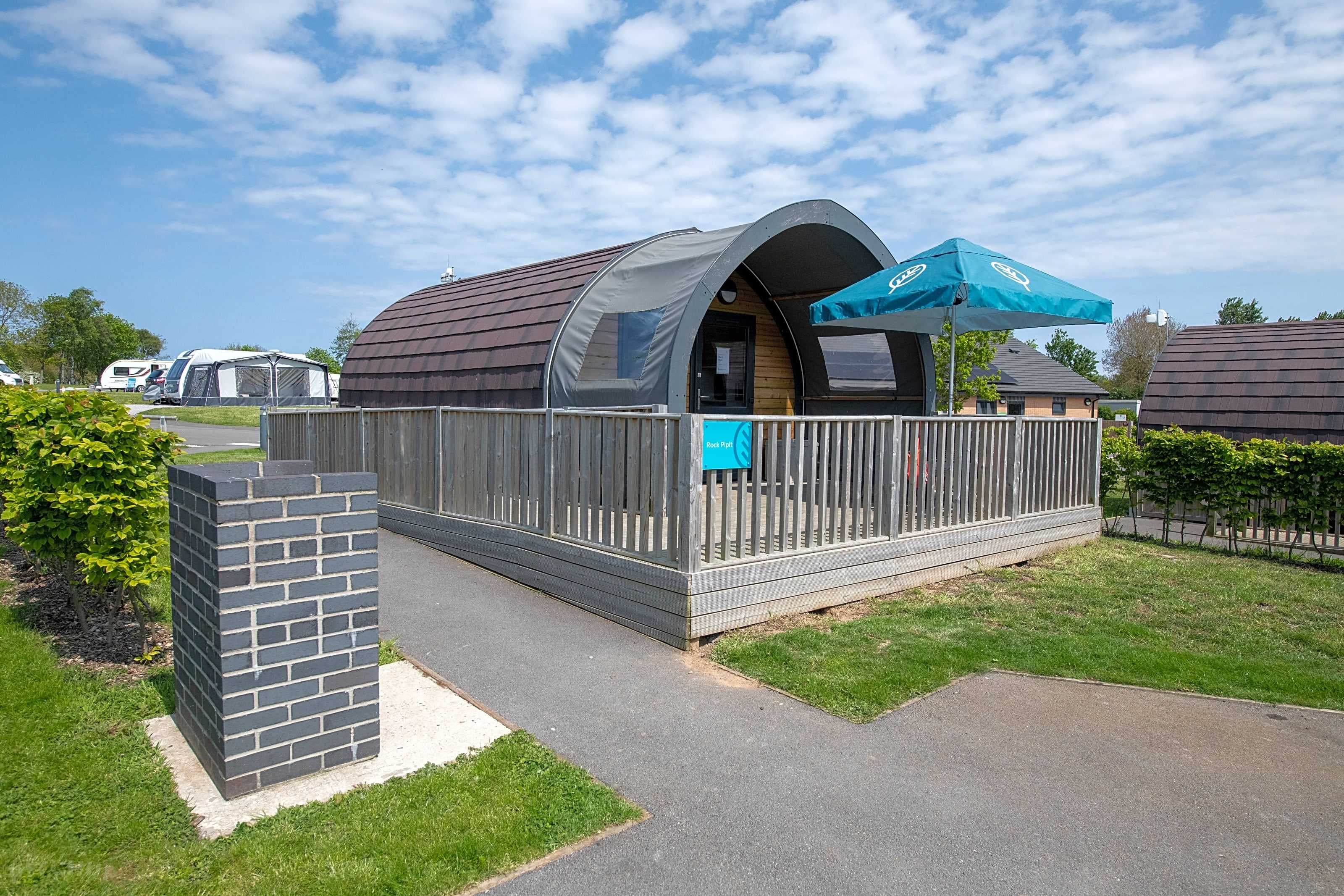 A universally accessible camping pod, with access ramp and blue umbrellas at the seating area